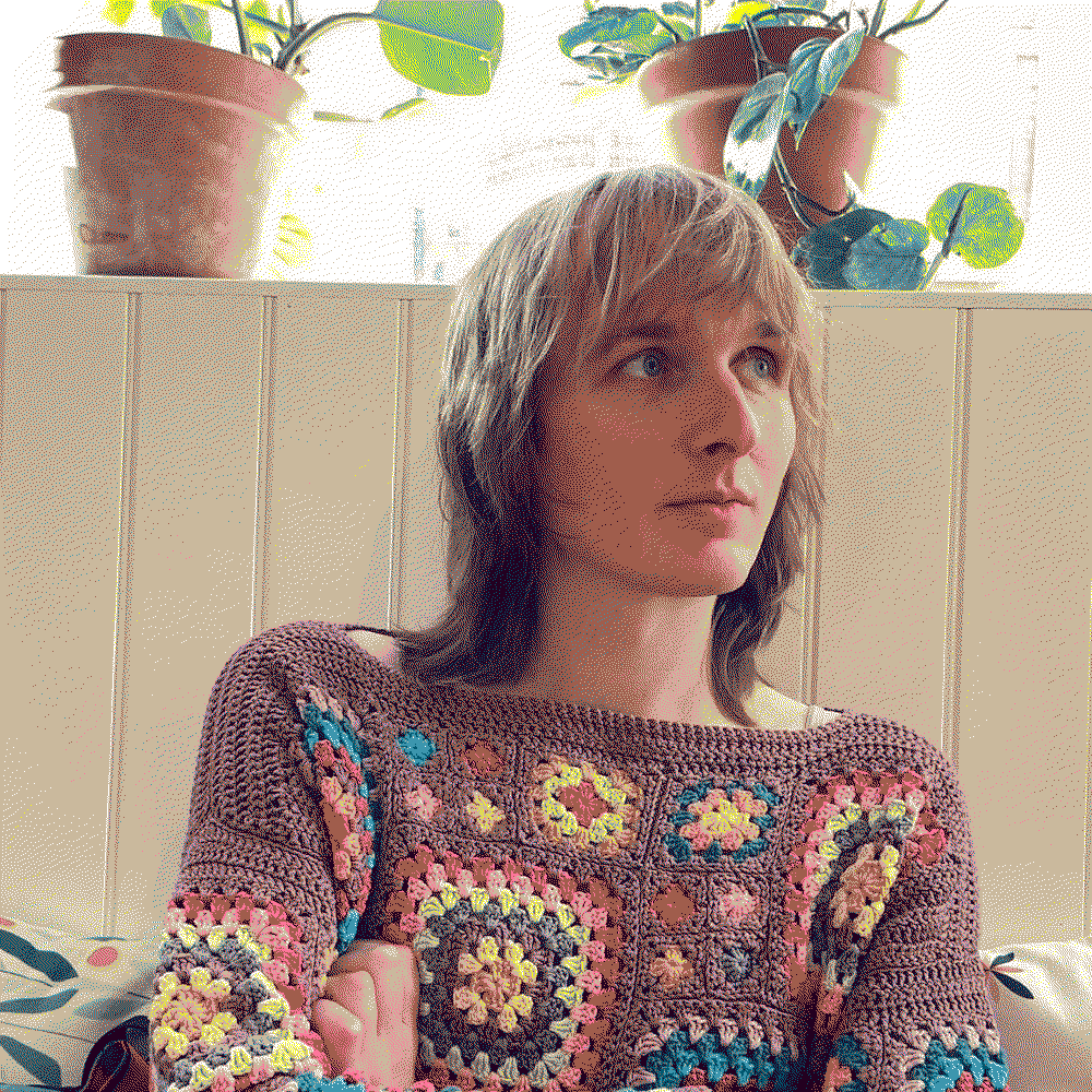 A shot of me with a fairly neutral if contented expression, folding my arms and looking off to the side and up a little. I'm sat on wooden-backed bench seating in a café. There are cushions to my sides and two plants on high window sills behind me. It's quite bright, but my portrait is clear. I'm white and have just-past-shoulder-length hair with a fringe. I'm wearing a knitted acrylic sweater, made by my mum. It's a base of purple with colourful patterned squares of differing sizes. Natural light is shining on one side of my face, from the direction I'm looking. The image is dithered, making the colour palette artistically muted